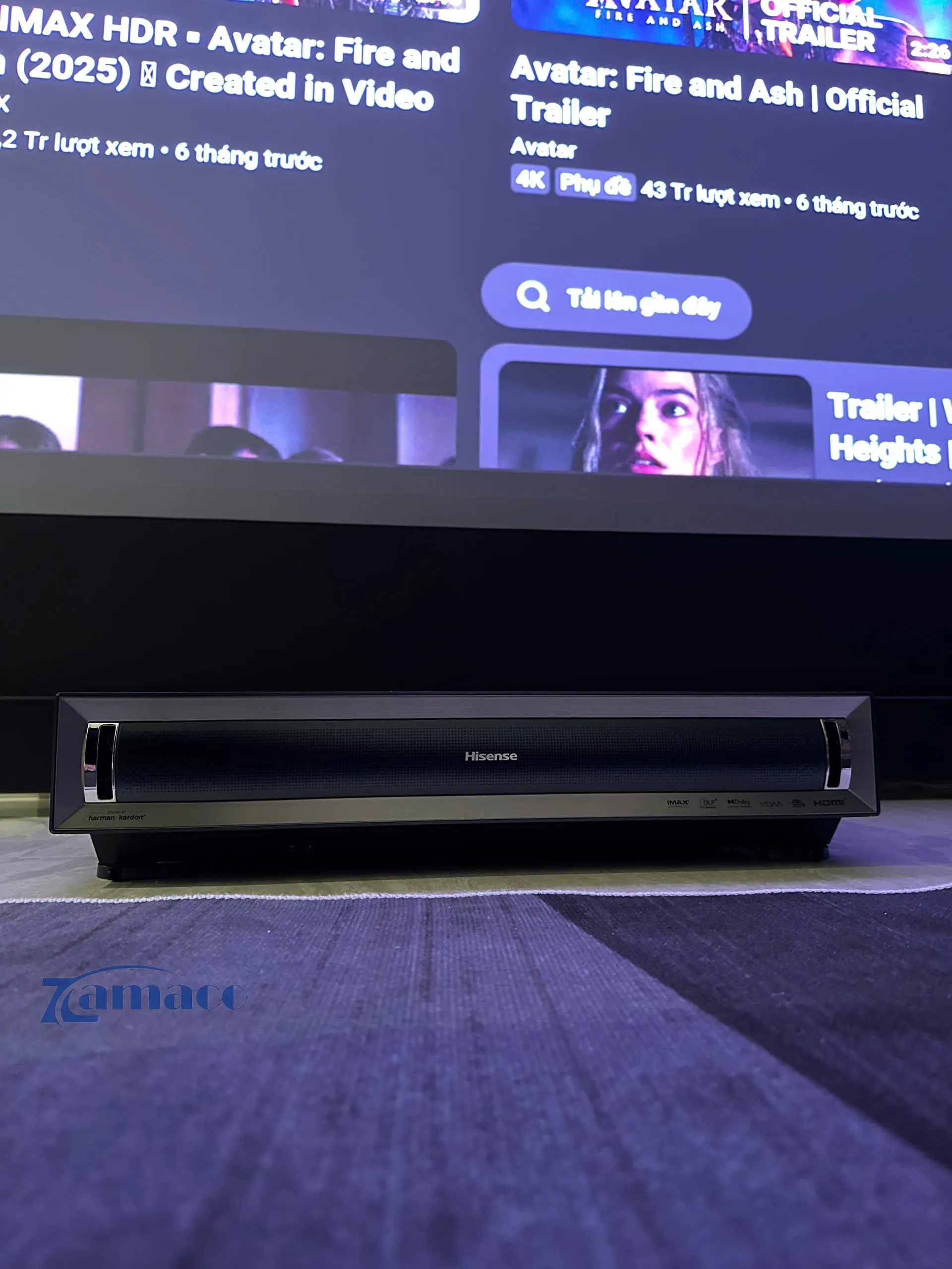 Máy chiếu siêu gần Hisense PX3 Pro 4K UST Laser Projector được đặt phía trước màn chiếu trong không gian phòng khách, thiết kế kim loại hiện đại với loa tích hợp phía trước, bố trí gọn gàng trên mặt sàn.