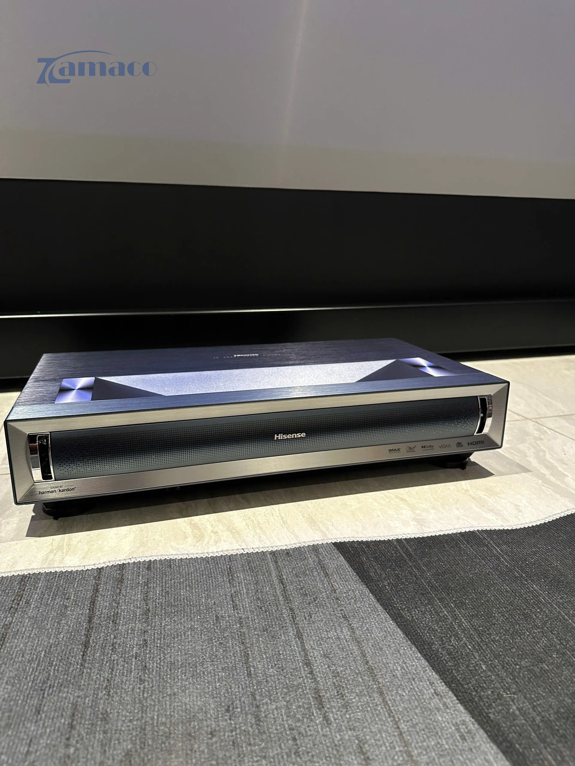 Máy chiếu siêu gần Hisense PX3 Pro 4K UST Laser Projector được đặt phía trước màn chiếu, thiết kế kim loại hiện đại và bố trí gọn gàng trên mặt sàn.