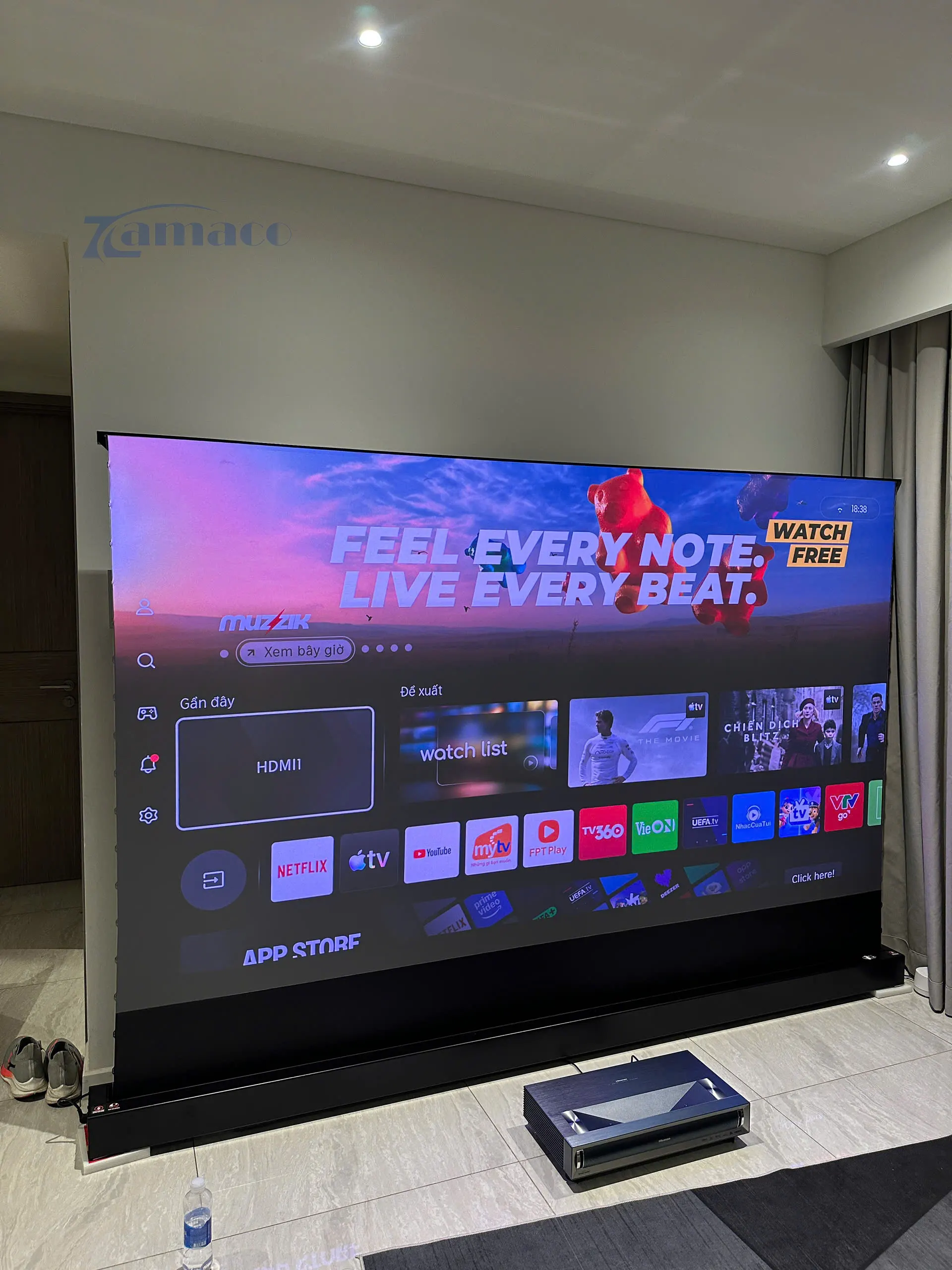 Máy chiếu siêu gần Hisense PX3 Pro 4K đặt dưới sàn phía trước màn chiếu Vividstorm VSDSTUST120H Floor Rising ALR Screen, trình chiếu giao diện Smart TV với hình ảnh lớn và sắc nét.