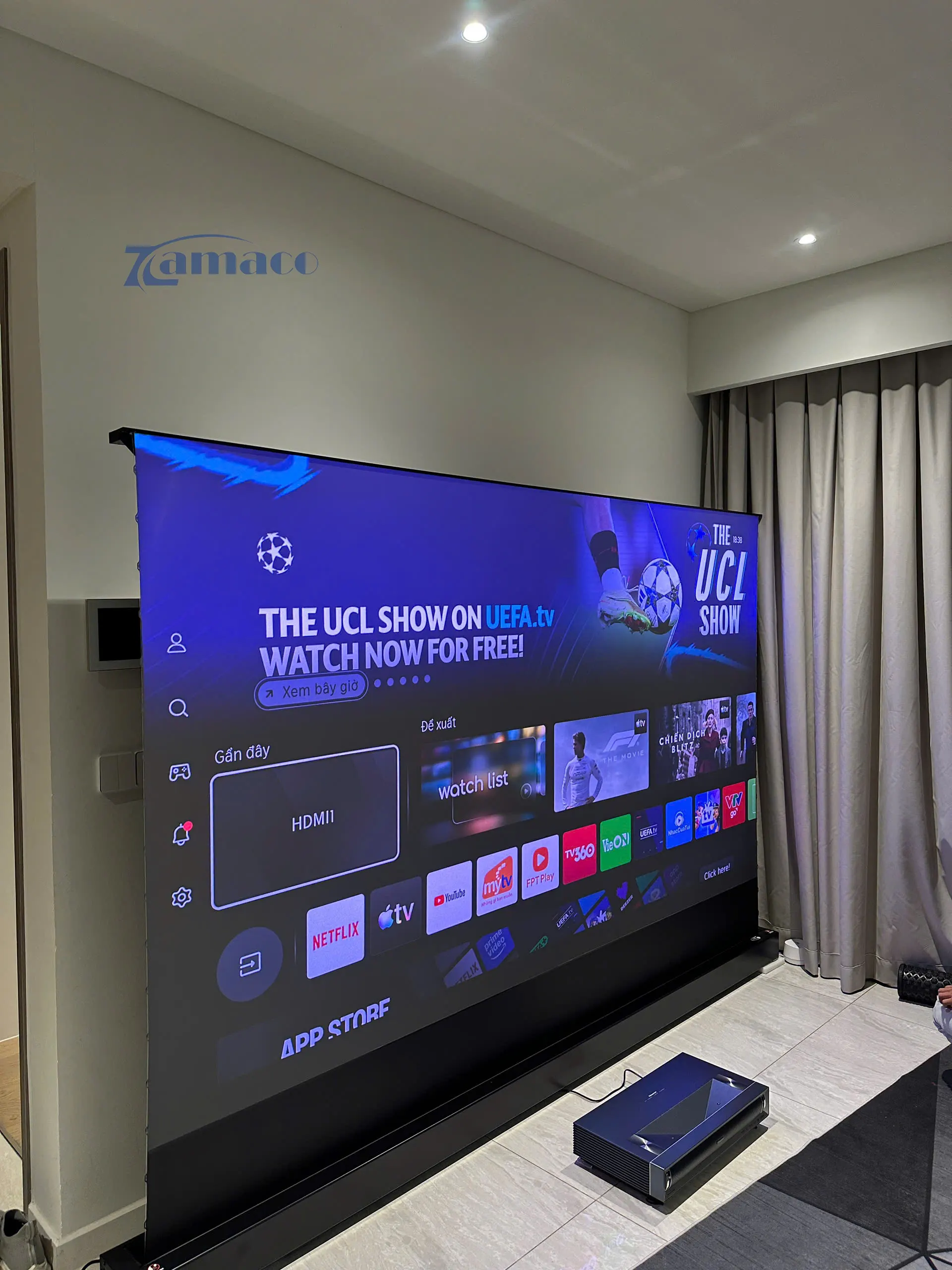 Máy chiếu siêu gần Hisense PX3 Pro 4K đặt dưới sàn phía trước màn chiếu Vividstorm VSDSTUST120H Floor Rising ALR Screen, trình chiếu giao diện Smart TV với hình ảnh lớn và sắc nét.